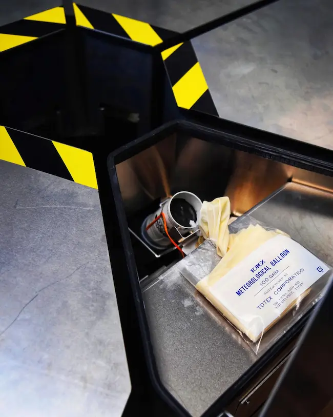 An open launch case with yellow-and-black caution striping, holding a seeding flare canister with its ignition wire, next to a sealed, unopened meteorological balloon package