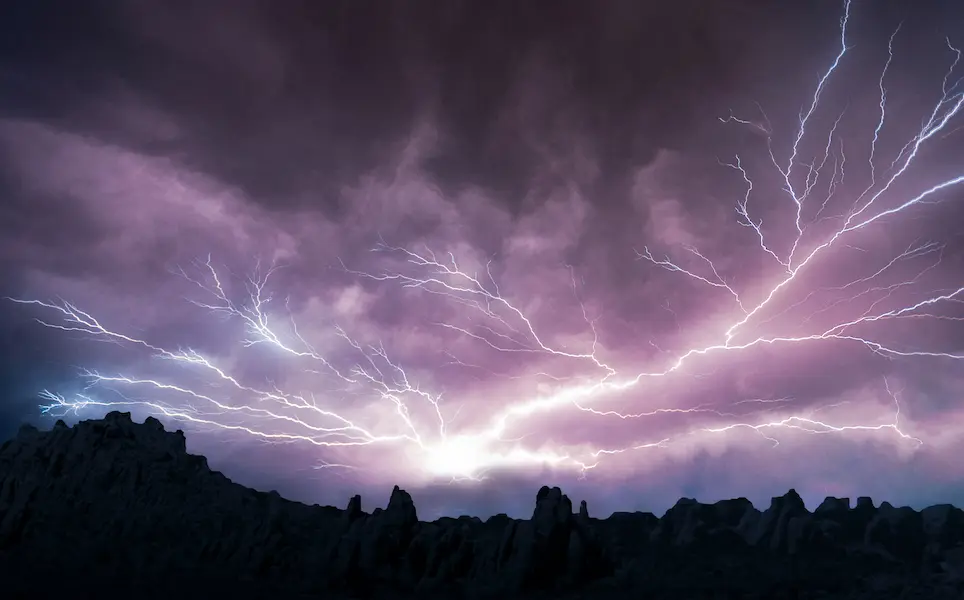Branching lightning bolts spreading across a dark purple storm sky above a silhouetted rocky landscape