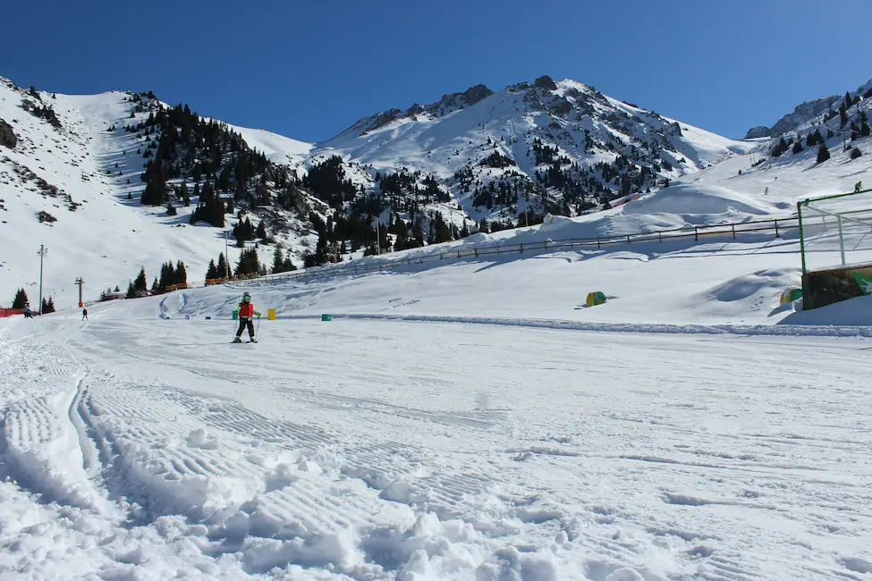 A skier descends a groomed snowy slope at a ski resort, with snow-capped mountains and a chairlift in the background